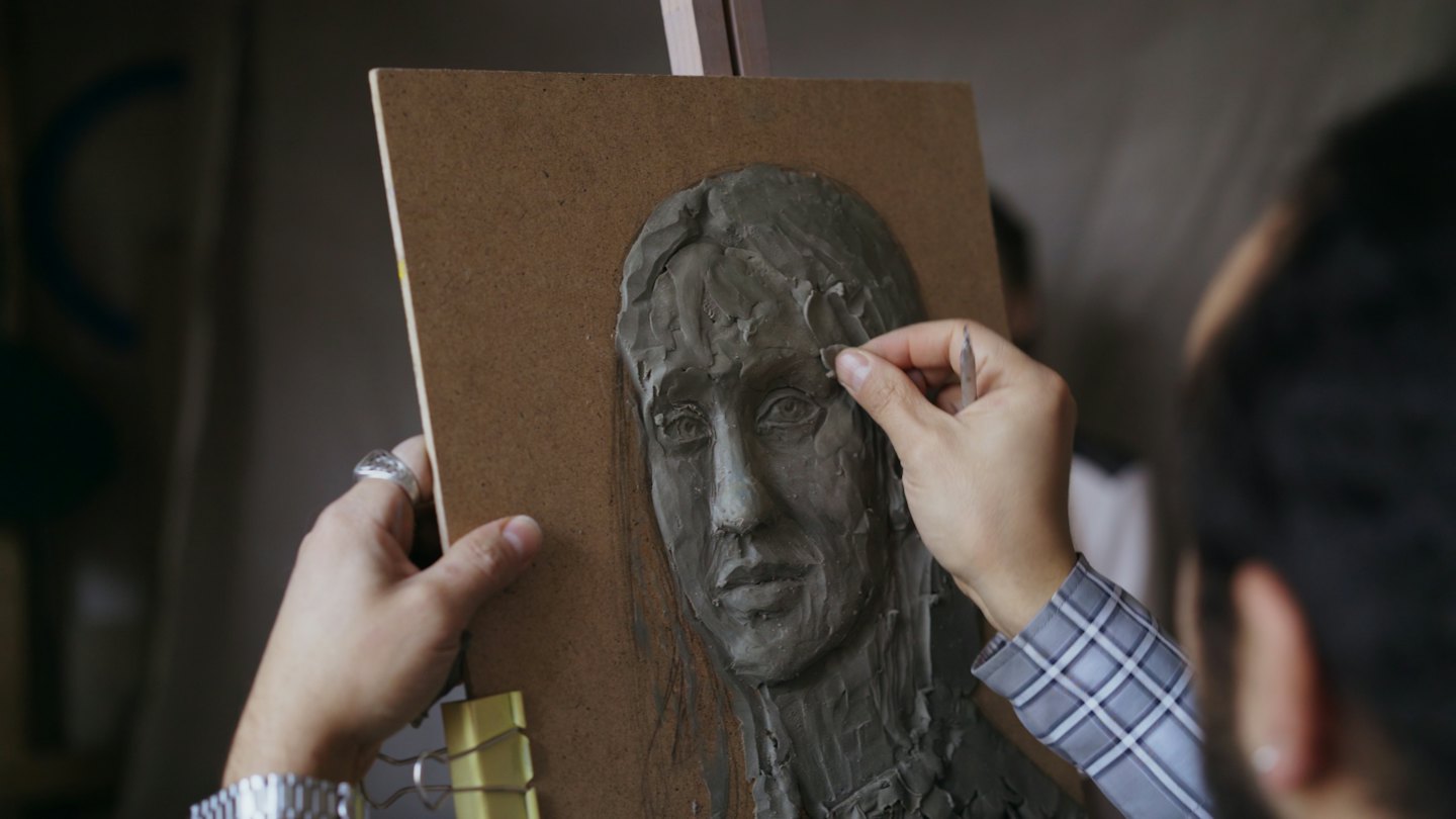 Hands sculpt a clay human face with a small tool.