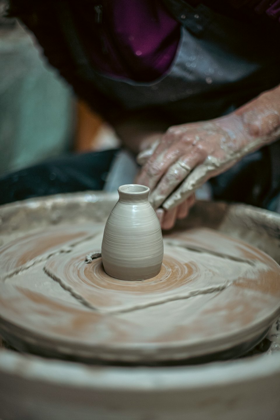 Hands covered in clay shaping a small vase on a pottery wheel.